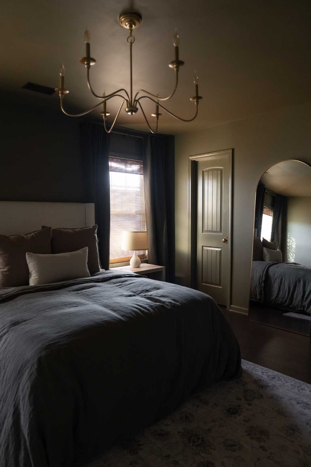 Moody bedroom with brass chandelier, dark linen bedding, arched floor mirror, and warm window glow